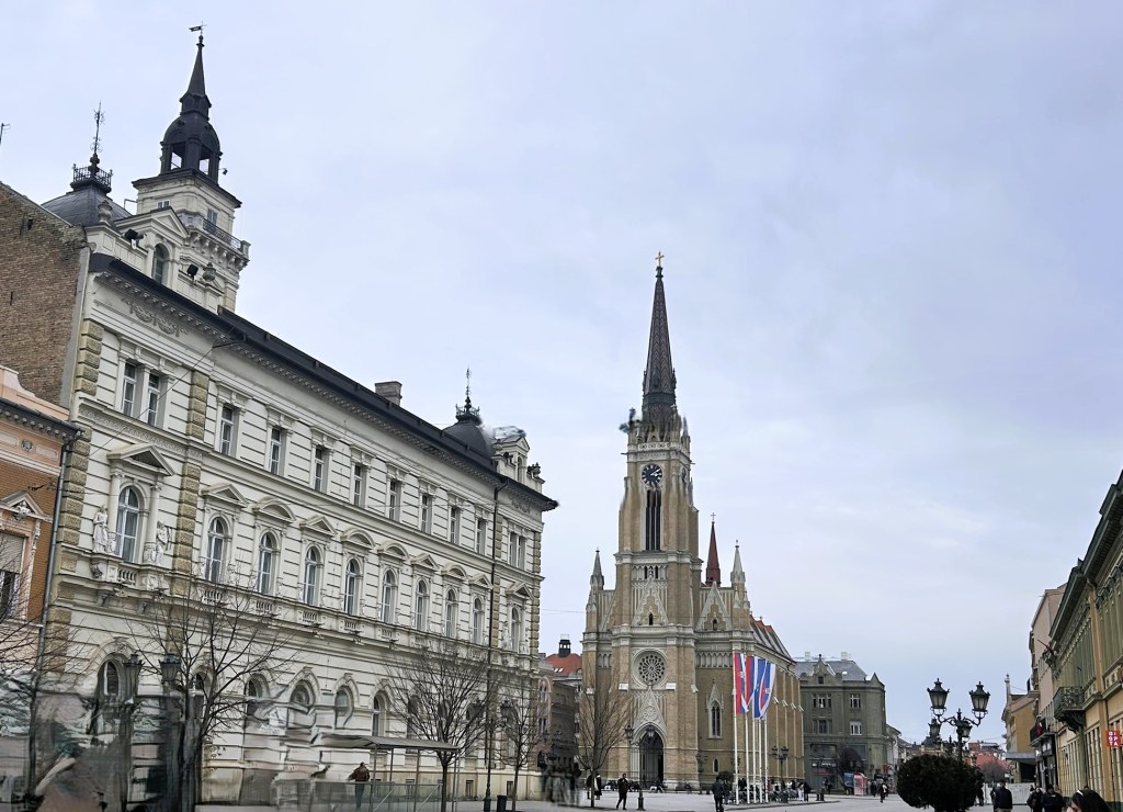 View of a historic square with ornate buildings, including a tall clock tower and decorative architecture, under a cloudy sky.