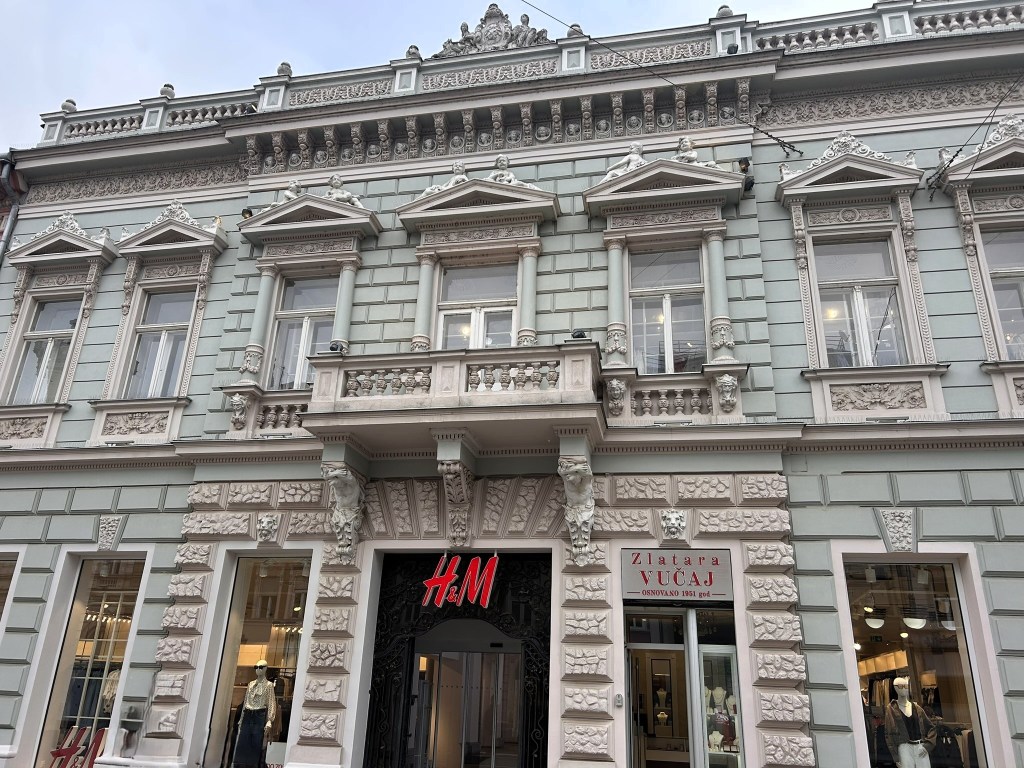 Facade of a historic building featuring elaborate architectural details, with an H&M entrance and a sign for a jewellery shop.