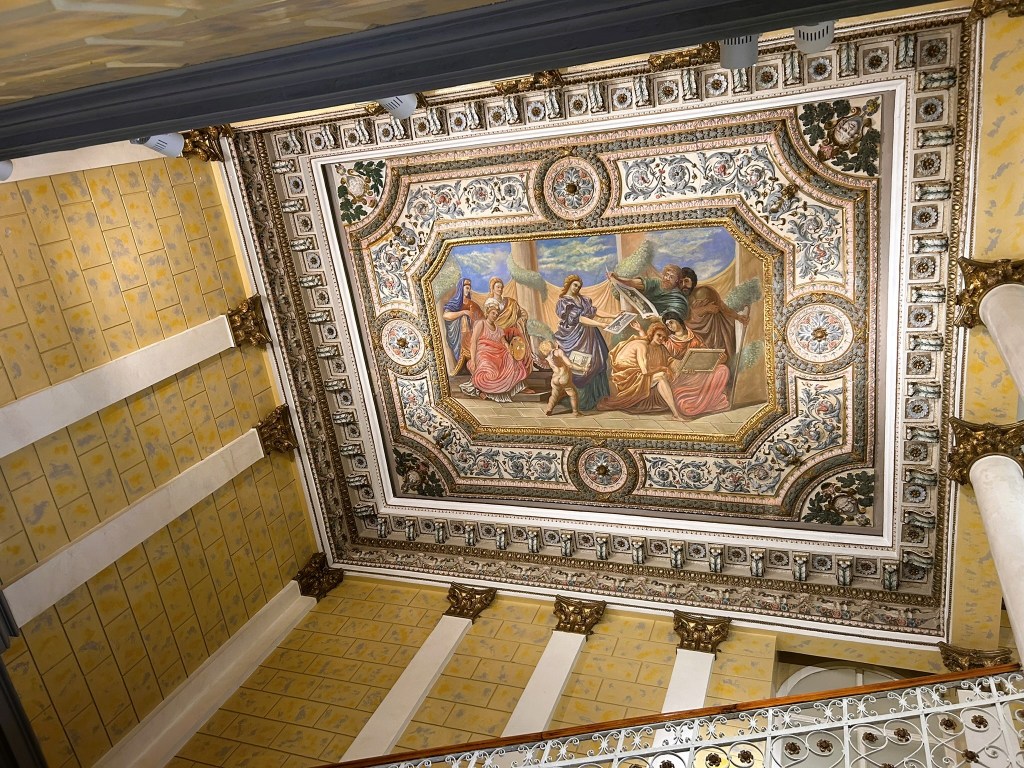 Decorative ceiling featuring a painted scene of figures, surrounded by intricate patterns and gold detailing, viewed from below.