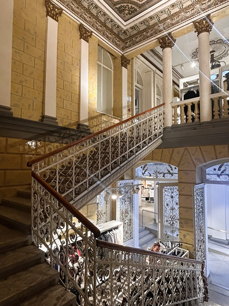 An ornate staircase with intricate wrought iron railings, leading to an upper level in a beautifully designed building featuring patterned ceilings and large windows.