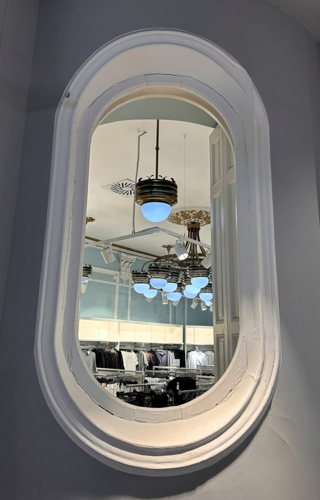A decorative oval mirror reflecting a well-lit shop interior featuring clothing racks and stylish pendant lights.