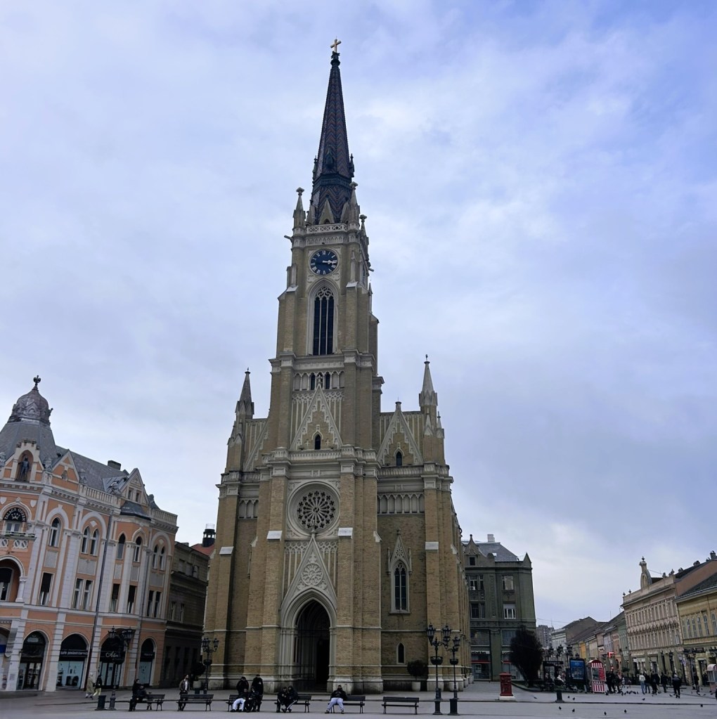 A tall church with a prominent spire and clock tower, located in an urban square with historic buildings surrounding it, under a cloudy sky.