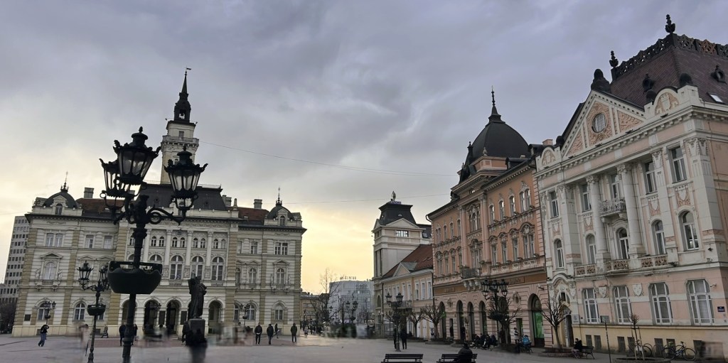 A city square featuring historical buildings with ornate architecture, a statue in the centre, and lampposts, under a cloudy sky.