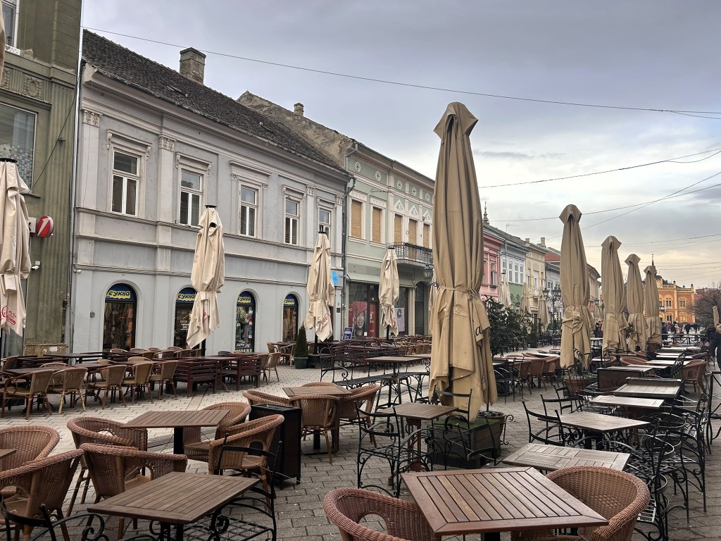 An outdoor cafe with numerous empty tables and chairs, shaded by large umbrellas, set in a European-style square with pastel-coloured buildings in the background.