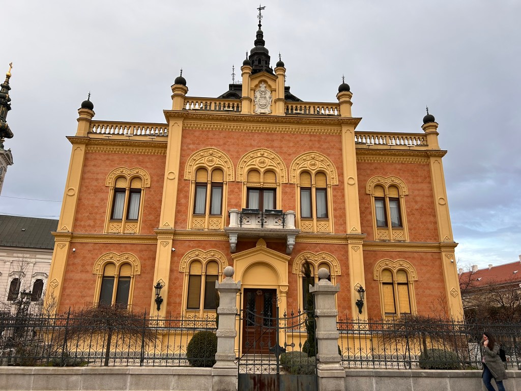 A large historic building with ornate yellow-orange façade and intricate architectural details, featuring multiple windows and a decorative roof, surrounded by a wrought iron fence and manicured greenery.