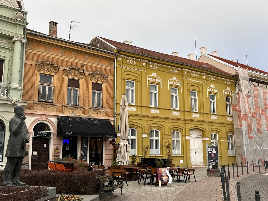 A street view of colourful historic buildings, featuring a yellow facade and a statue of a man in the foreground. Outdoor seating with tables and an umbrella is visible in front of a restaurant.
