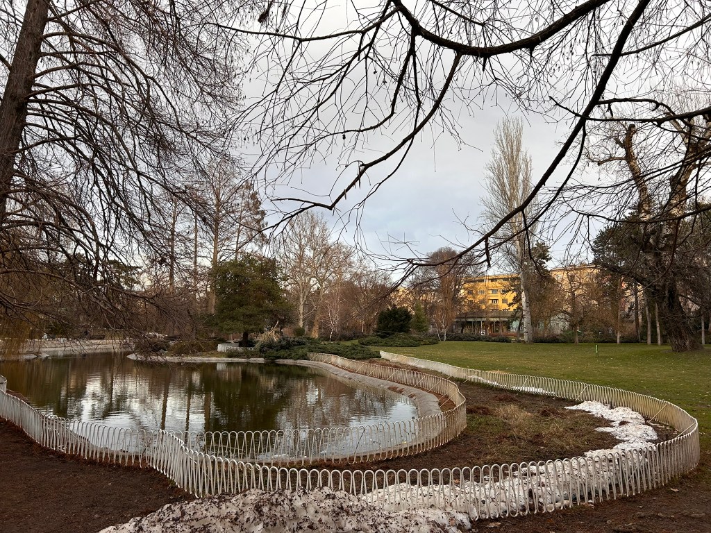 A serene park scene featuring a pond surrounded by a white fence, with bare trees in the foreground and a grassy area in the background under a cloudy sky.