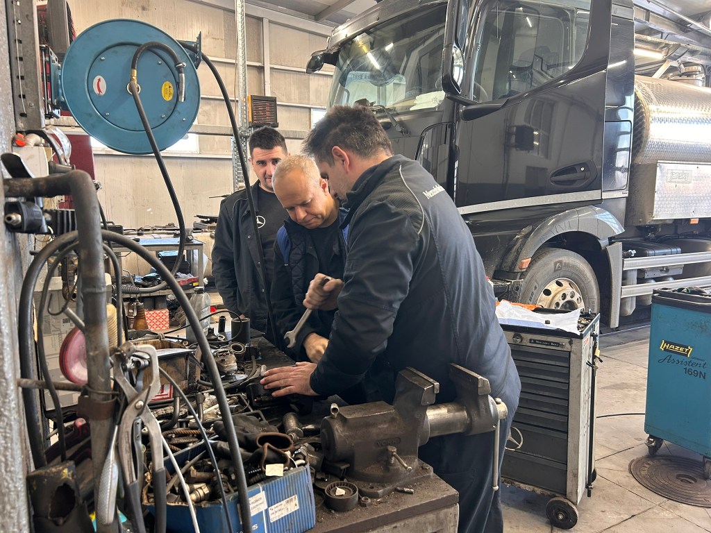 Three mechanics examining and repairing machinery in a garage, with a large truck partially visible in the background.