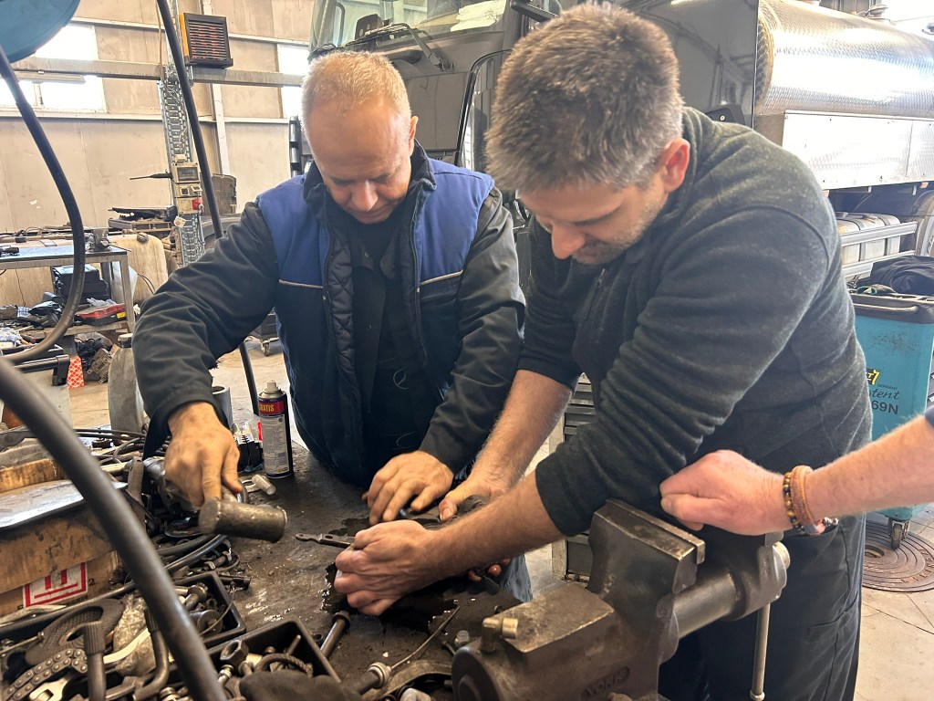 Two mechanics working together on a vehicle part in a workshop, using tools at a cluttered workbench.