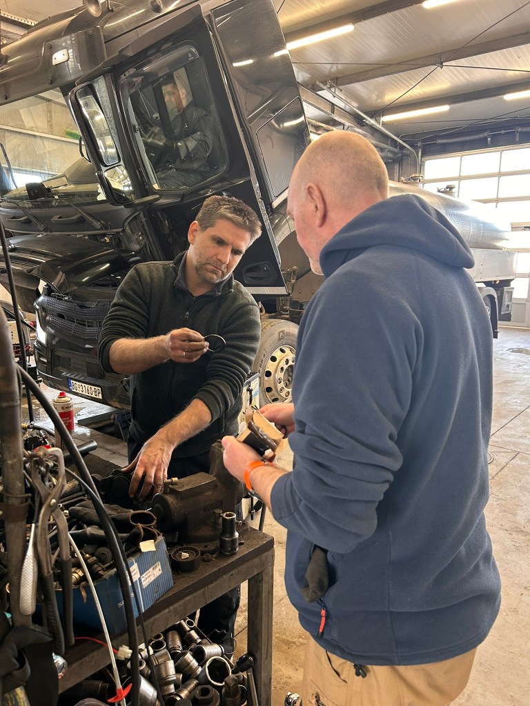 Two mechanics discussing tools and repairs in a workshop, with a black truck in the background.