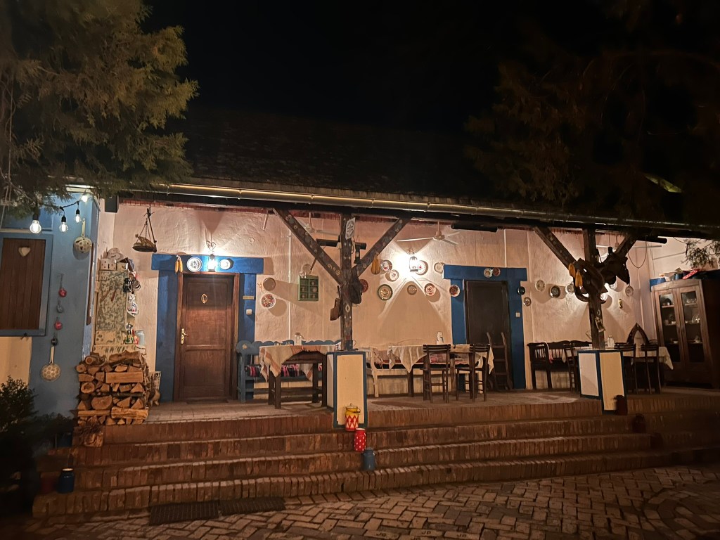 Exterior view of a charming restaurant at night, featuring wooden doors, blue and white walls, and decorative plates. Tables and chairs are set outside under soft lighting.