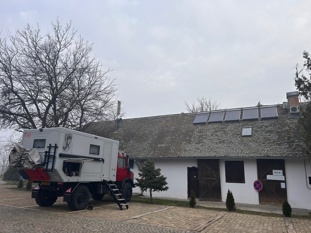 A large overland vehicle parked in front of a traditional building with a sloped roof, featuring solar panels. The vehicle has a unique design, and there are trees and a sign visible nearby.
