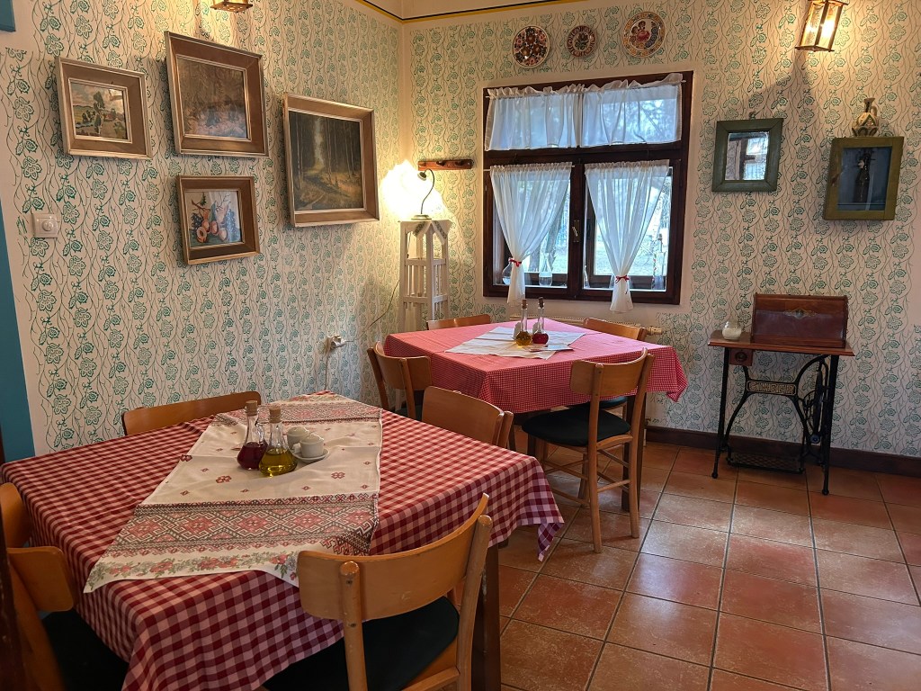 Interior of a quaint restaurant featuring two wooden tables with checkered tablecloths, decorated walls with framed paintings, and a window with white curtains.