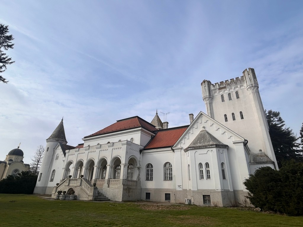A large white castle with a red roof, featuring two towers, surrounded by green grass and trees, under a cloudy sky.