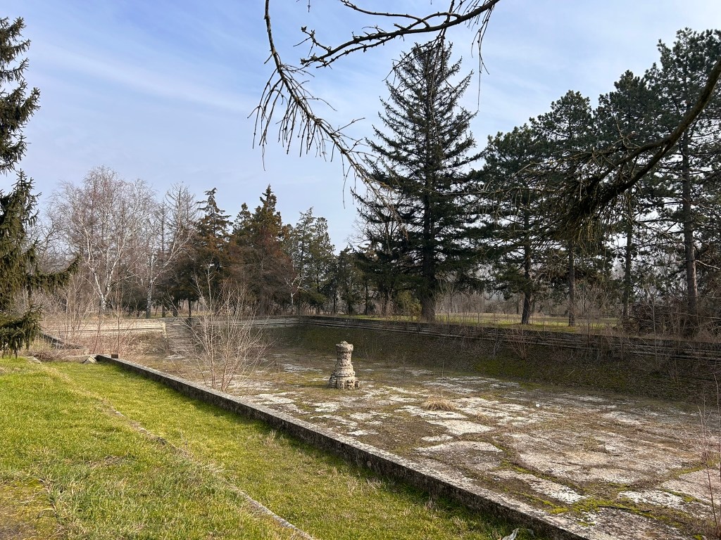 An overgrown stone platform surrounded by trees, with a stone structure in the centre and a vast expanse of grass and bare ground.