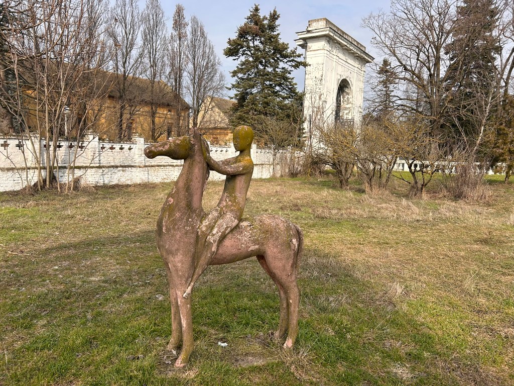A sculpture of a figure riding a horse, set in a grassy area with trees and a historic structure in the background.
