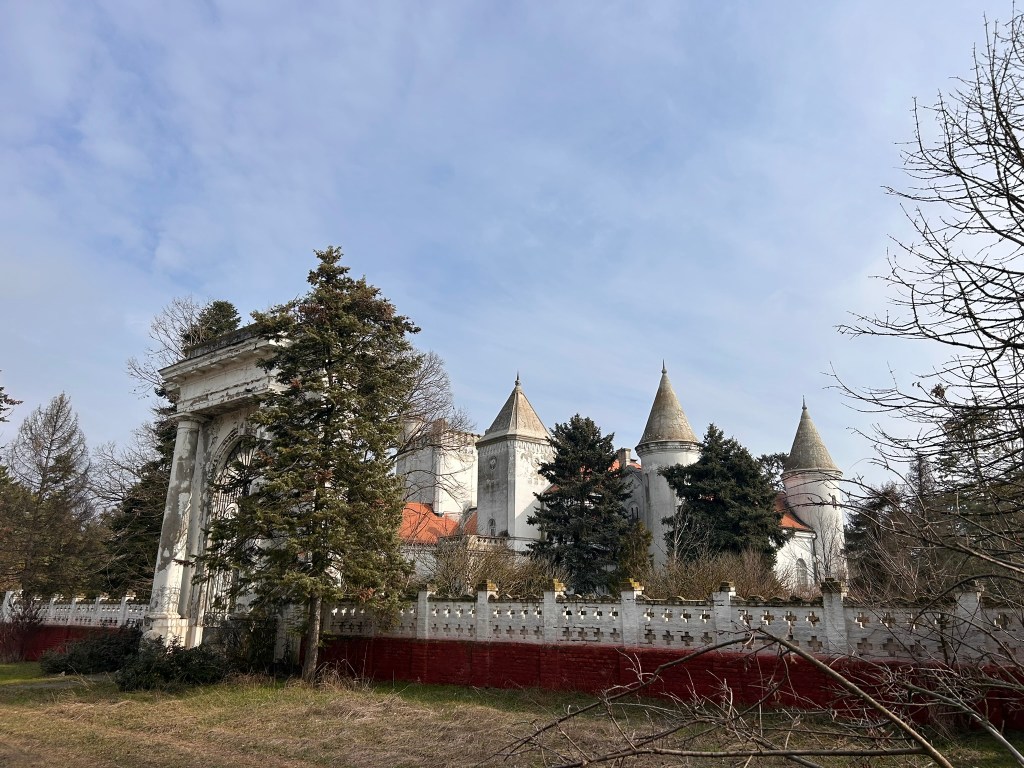 A historic castle or manor set against a cloudy sky, partially obscured by trees and surrounded by a decorative wall.