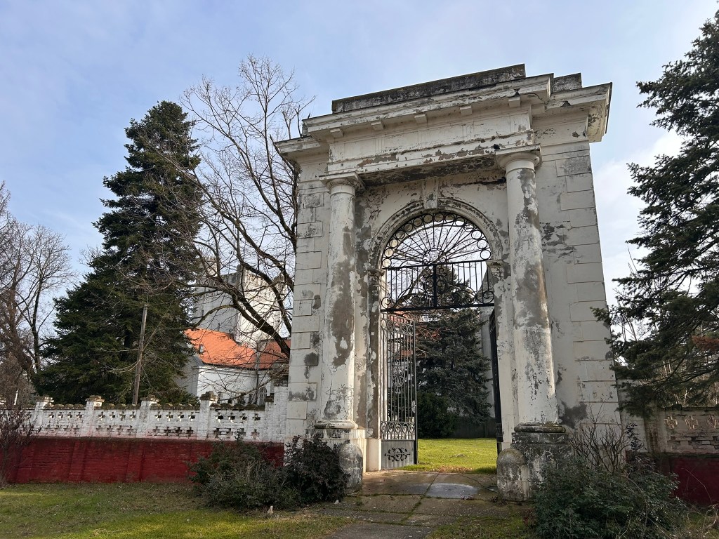 An ornate, weathered stone archway with a wrought iron gate, set against a background of trees and a grassy area.