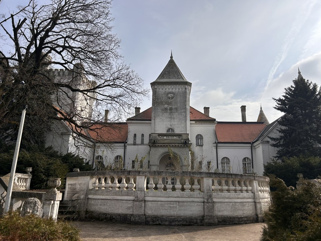 A large, historic mansion with a tower, surrounded by trees and a stone wall, under a cloudy sky.