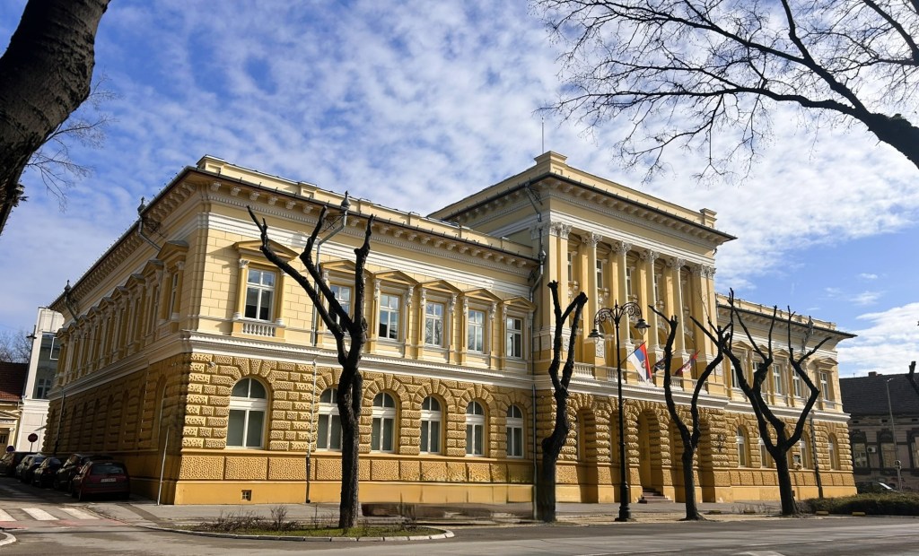 A historic yellow building with ornate architectural details and large windows, set against a partly cloudy sky. The front of the building features trees with bare branches and street lamps.