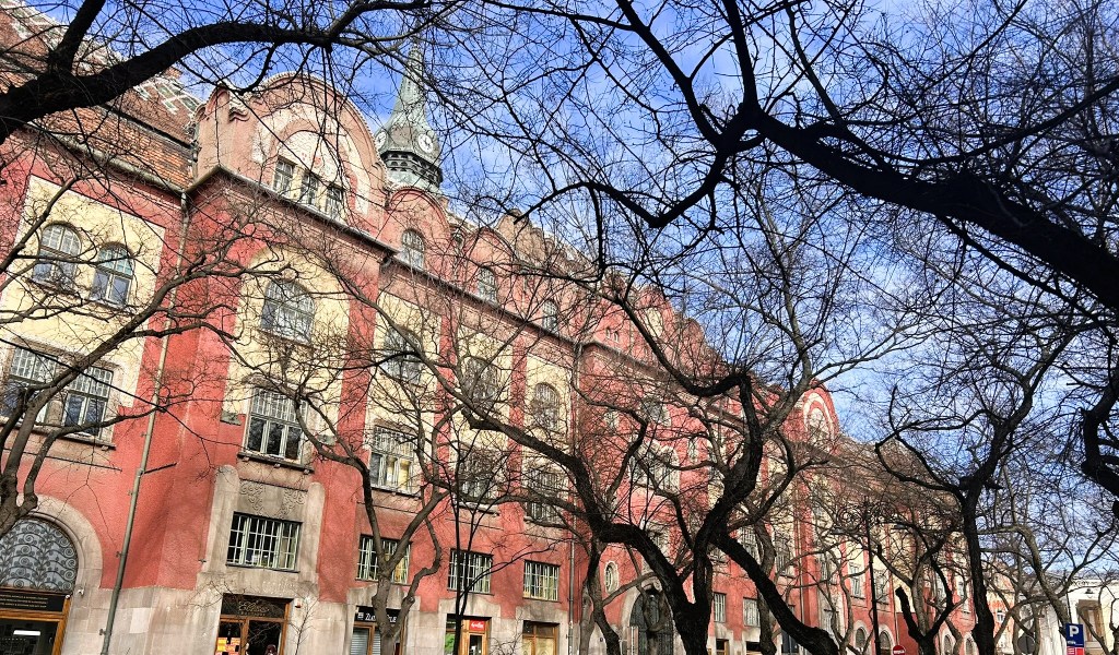 Historic building with a clock tower, partially obscured by bare tree branches, under a blue sky.