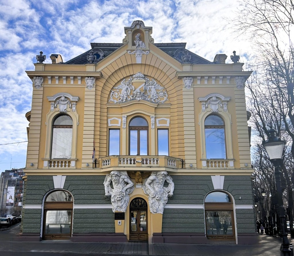 Ornately designed yellow and green building with sculptural elements, featuring large windows and intricate architectural details under a cloudy sky.
