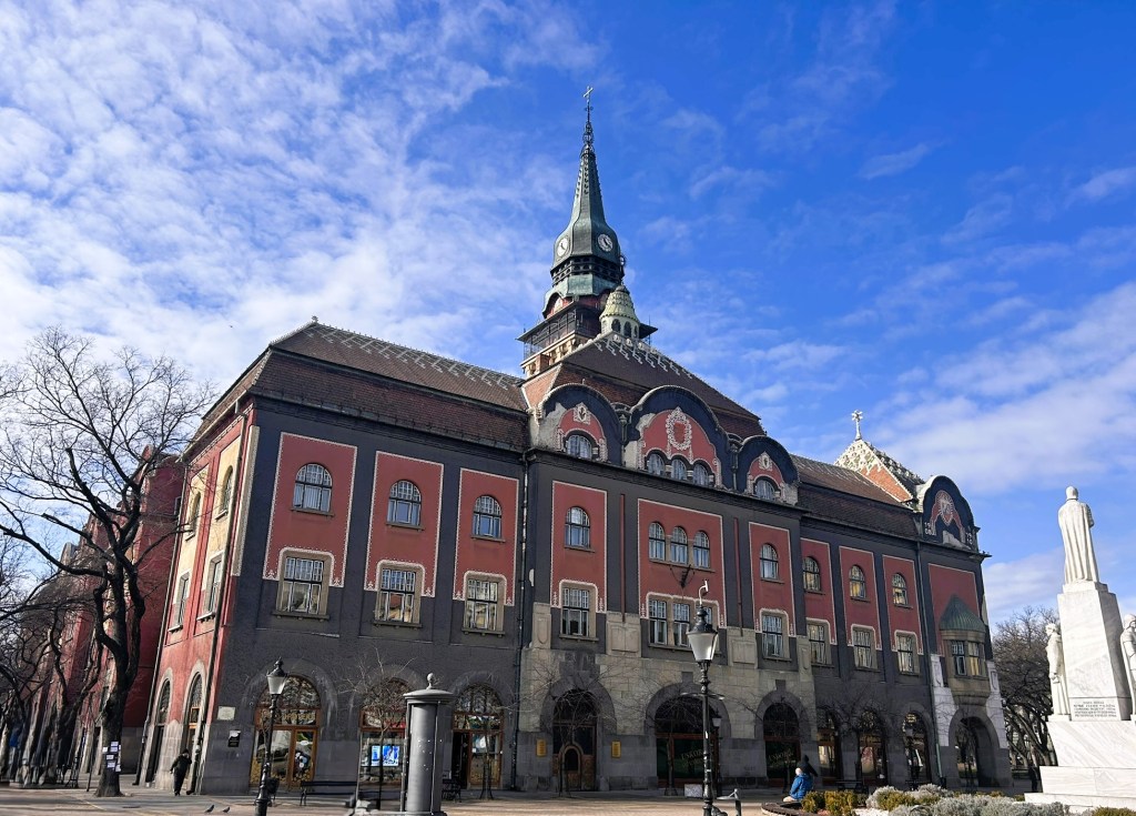 A historic building with a tall spire, featuring red and grey façades, and intricate architectural details, set against a clear blue sky.