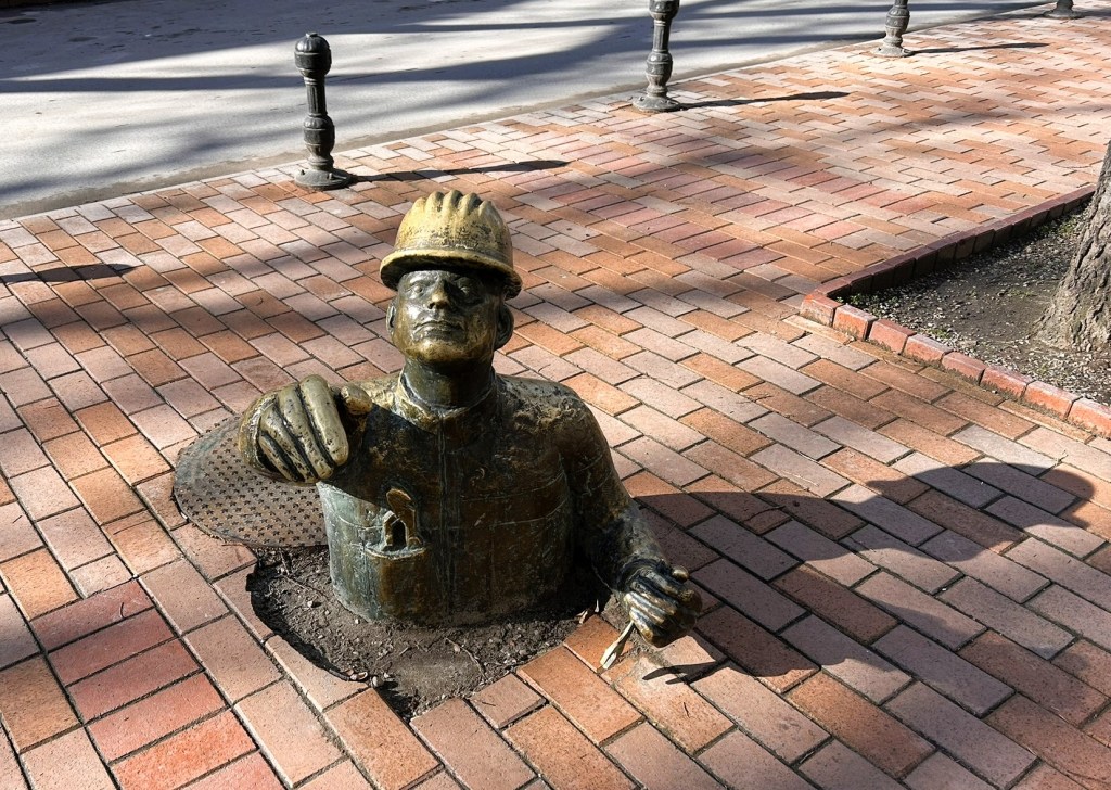 A bronze sculpture of a worker in a hard hat emerging from the ground, holding tools, situated on a brick pavement.