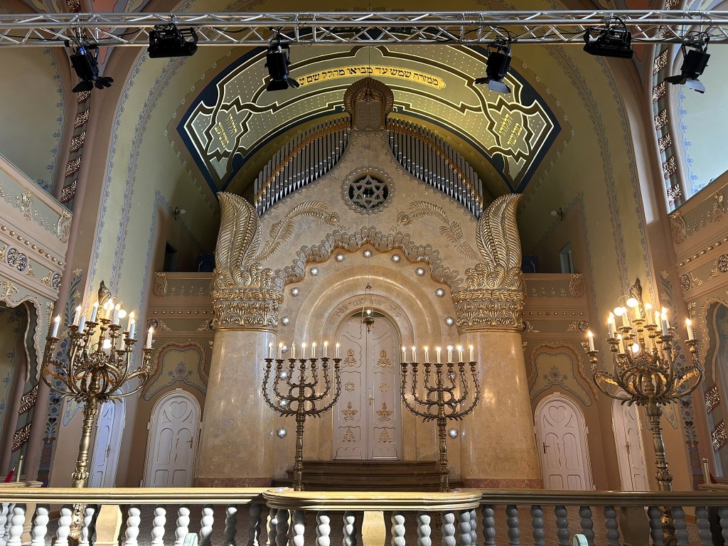 Interior view of a beautifully decorated synagogue, showcasing a grand bimah with ornate features, a large organ, and two elegant candelabras with lit candles.