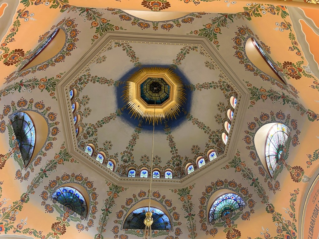 View of an ornate octagonal dome featuring intricate floral designs and stained glass windows, illuminated by a central light fixture.