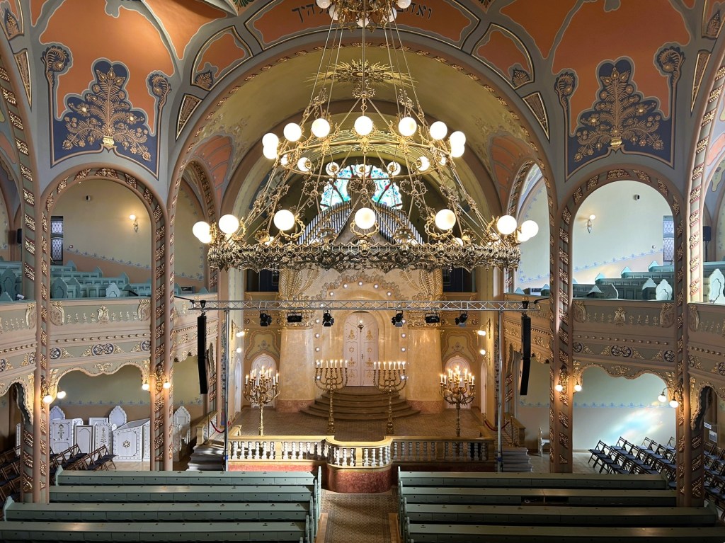 Interior view of a beautifully decorated synagogue featuring ornate ceilings, chandeliers, and rows of seating.