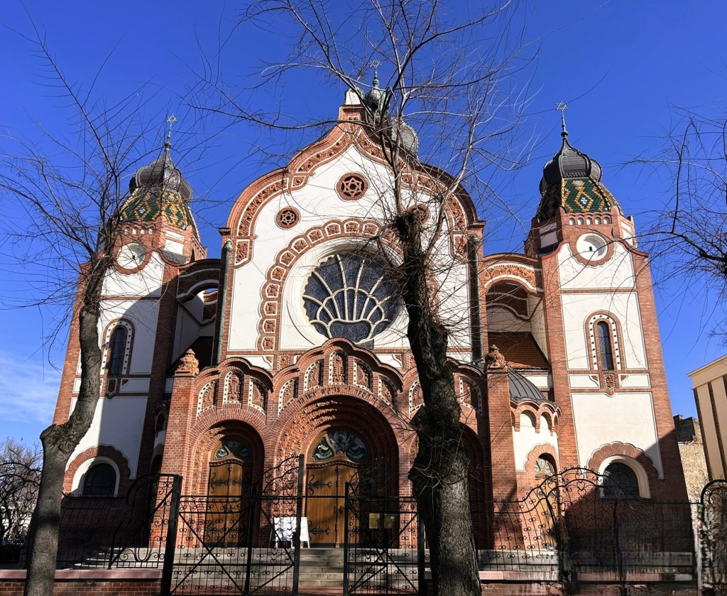 Front view of a decorative building with domes, intricate brickwork, and a large arched window against a clear blue sky.