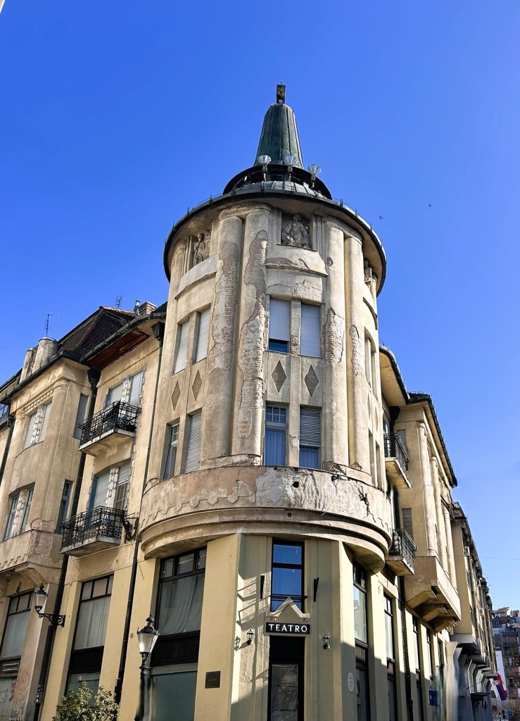 A historic building with a rounded corner, featuring a decorative facade and a pointed rooftop, illuminated against a clear blue sky. The ground floor displays a sign that reads 'Teatro'.