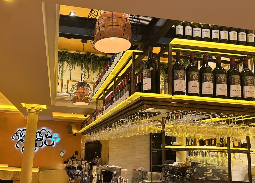 Interior of a stylish bar featuring illuminated shelves displaying wine bottles and glasses, with decorative pendant lights and an elegant atmosphere.
