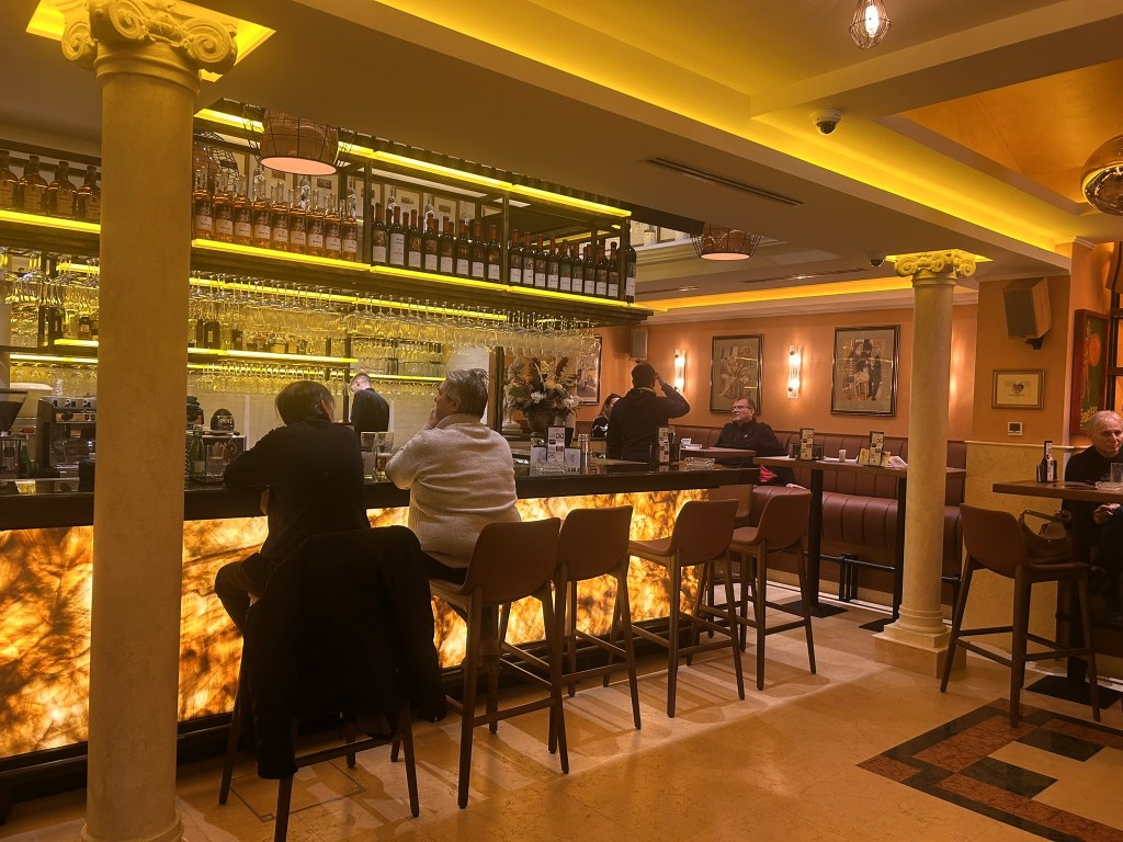 Interior of a stylish bar featuring a glowing backlit counter, patrons seated on high stools, and various bottles displayed on shelves behind the bar. Warm lighting and decorative art create a welcoming atmosphere.
