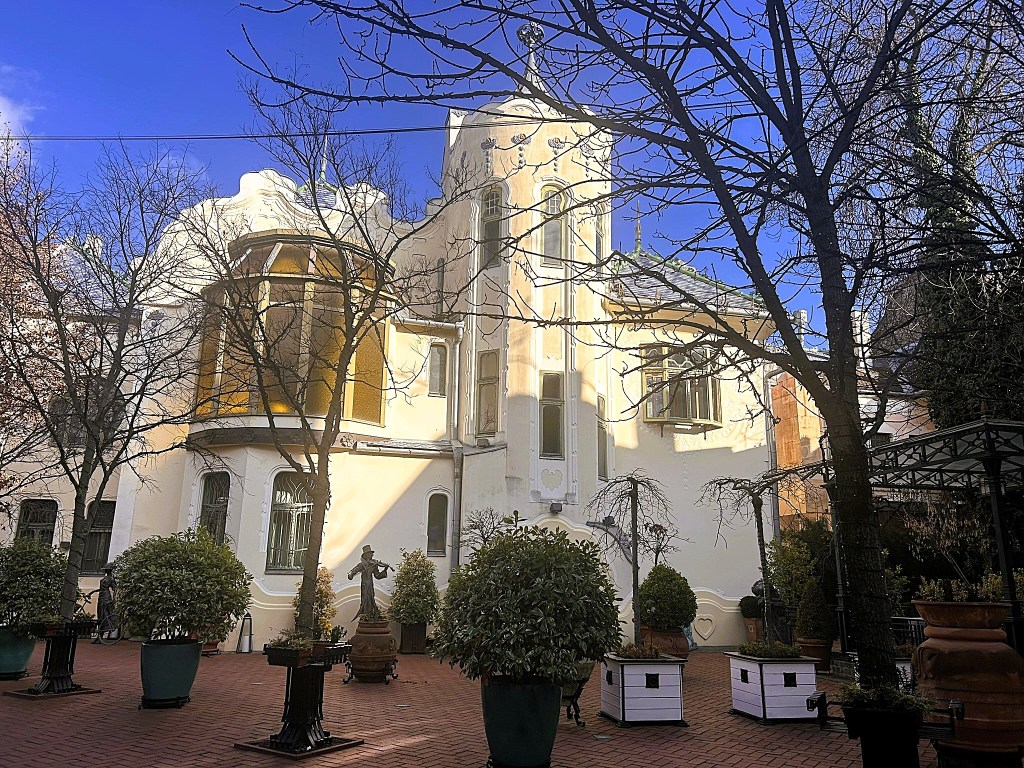A scenic view of a white architectural building with curved elements, surrounded by bare trees and planters in a courtyard. The sky is clear and blue, enhancing the tranquil atmosphere.