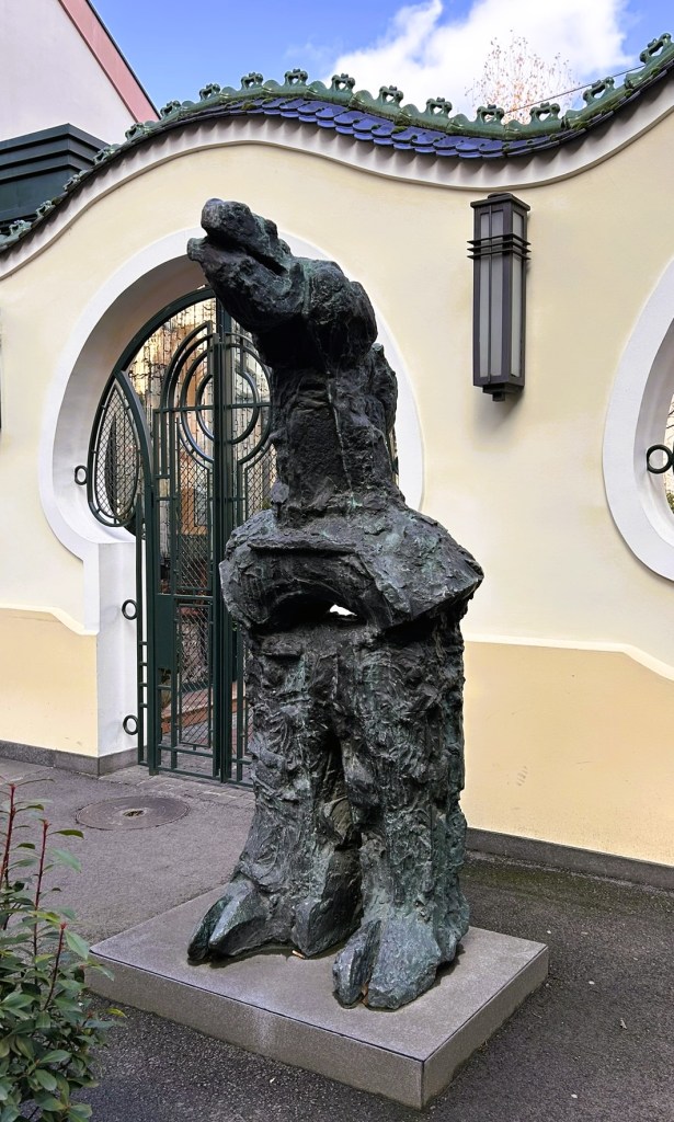 A bronze sculpture of a standing bear in an urban setting, positioned on a stone base, with a decorative wall and gate in the background.
