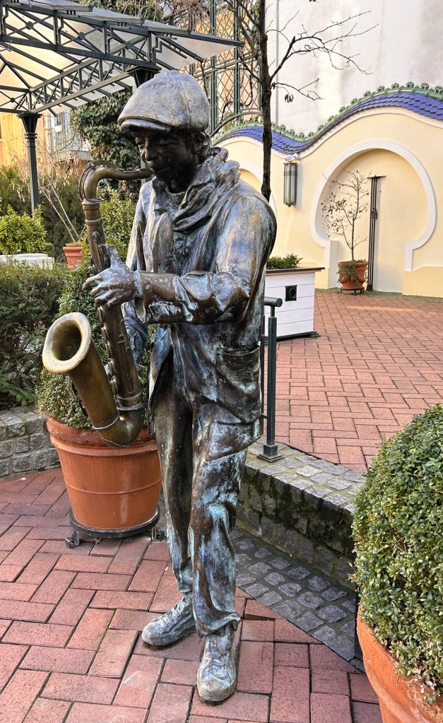 Bronze statue of a musician playing a saxophone, set in a garden with ornamental plants and brick pathways.