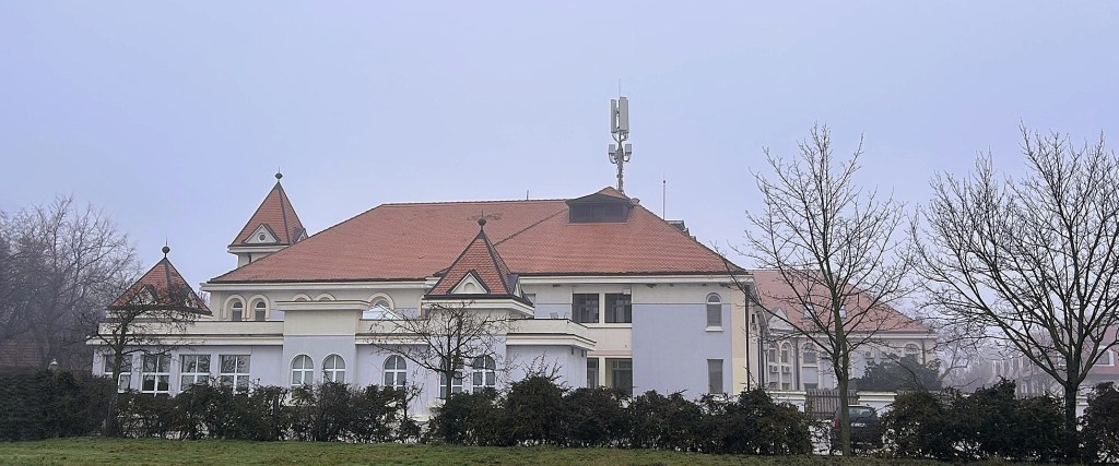 A large, two-storey building with decorative peaks on the roof, surrounded by trees and hedges, in a foggy environment.