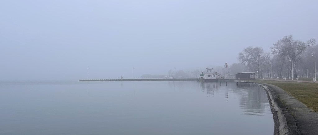 A misty lakeside view with calm waters and a distant pier, surrounded by trees and a foggy atmosphere.