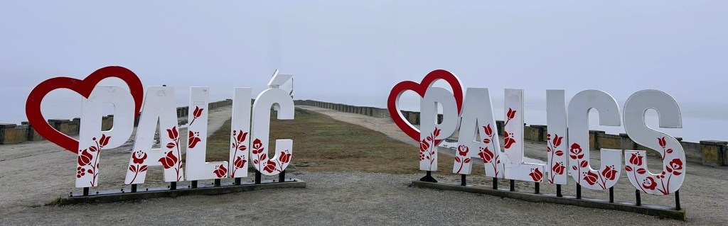 Large decorative sign featuring the text 'I love Palić' with floral designs, located by a pathway near a body of water, on a foggy day.