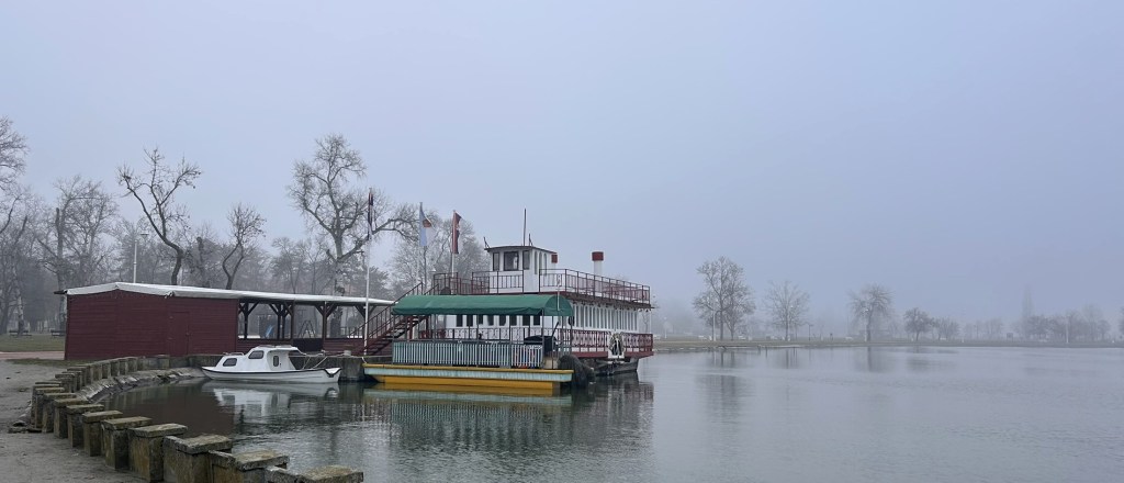 A misty landscape featuring a boat docked near a ferry-style building by a calm lake, surrounded by bare trees and a cloudy sky.