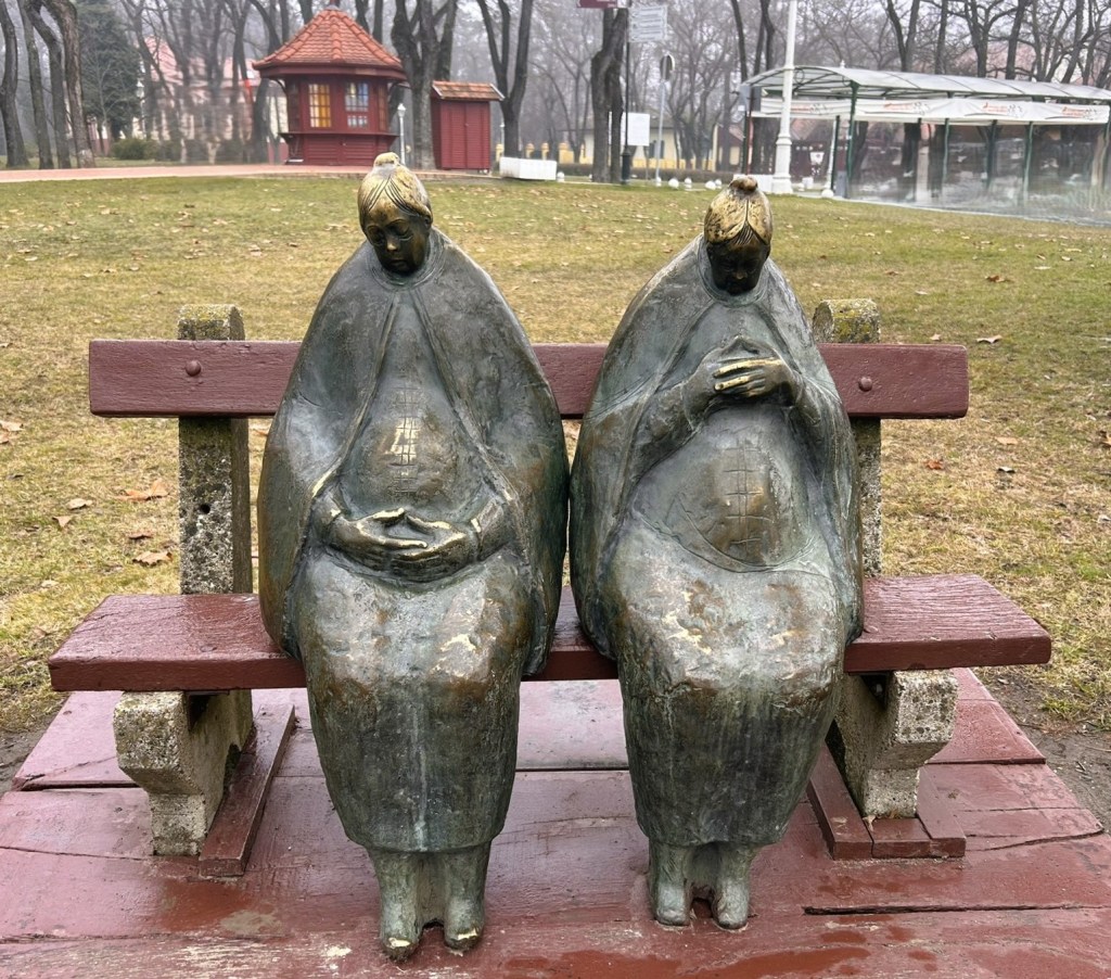 Two bronze statues of women in traditional clothing seated on a wooden bench in a park setting.