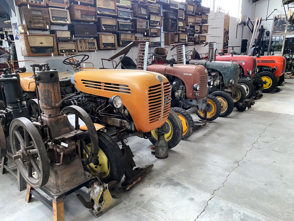 A row of vintage tractors displayed in a museum, showcasing various colours and designs against a backdrop of antique radios mounted on the wall.