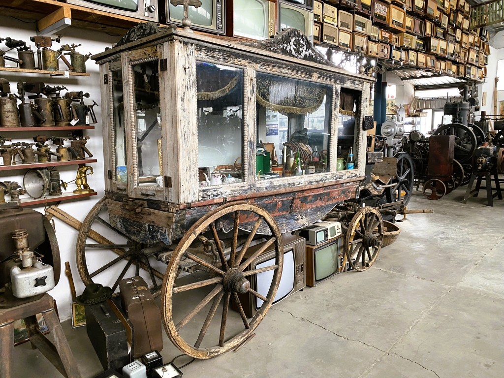 An antique wooden cart with glass panels on display in a museum filled with vintage items and machinery.