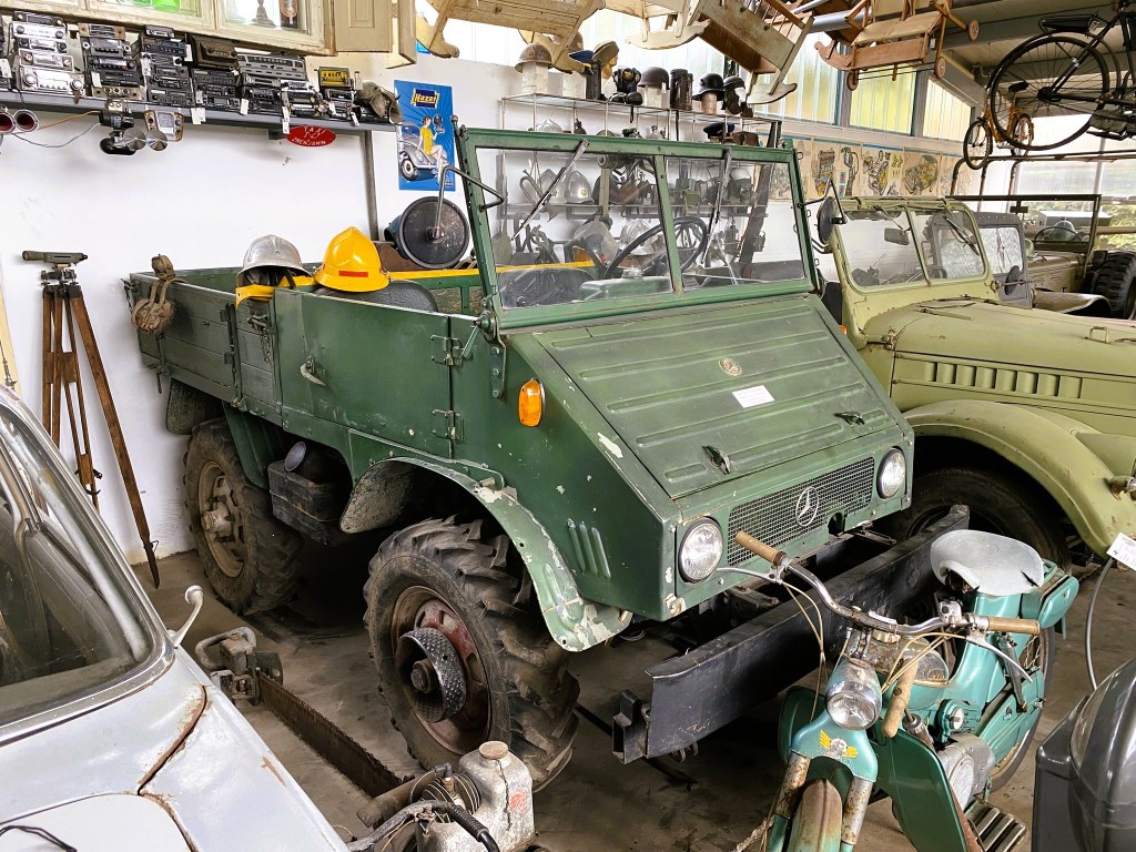 A vintage green military-style vehicle is parked inside a museum, surrounded by various old vehicles and memorabilia. The vehicle features a simple design with exposed wheels and equipment on the dashboard. A yellow hard hat and grey seat cushions are visible inside.