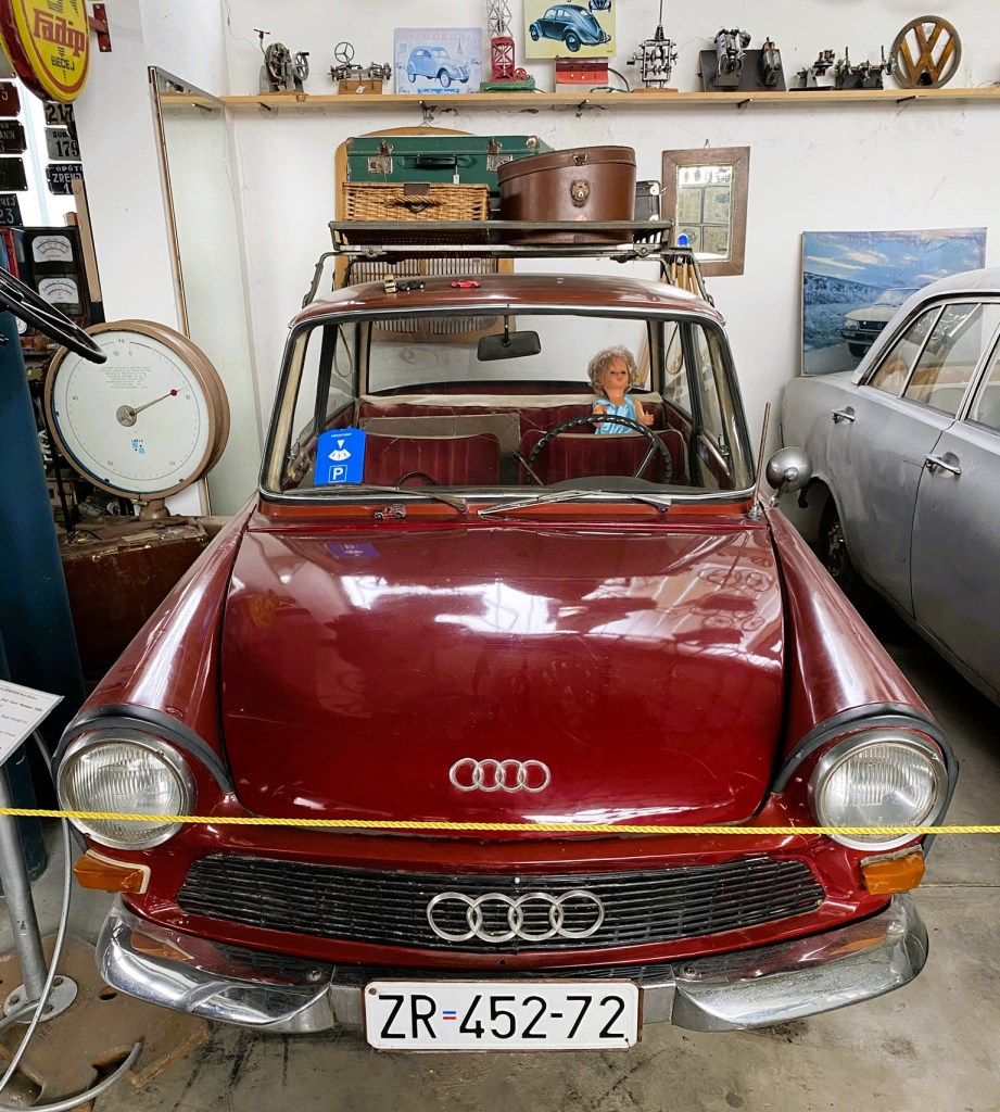 Vintage maroon Audi car displayed in a museum, featuring a doll in the driver's seat and various old memorabilia in the background.