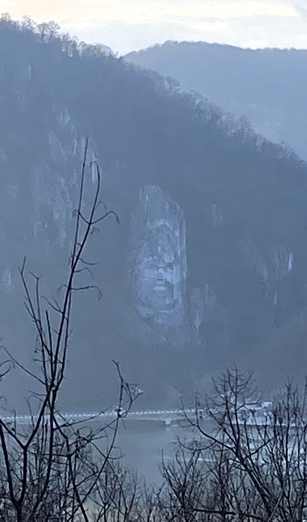 A misty view of a mountain with a large rock formation resembling a face, surrounded by bare trees and a river with a bridge in the foreground.