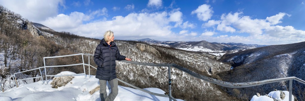 A person stands by a safety railing on a snowy mountain overlook, gazing at a panoramic view of rolling hills and valleys under a blue sky with fluffy clouds.