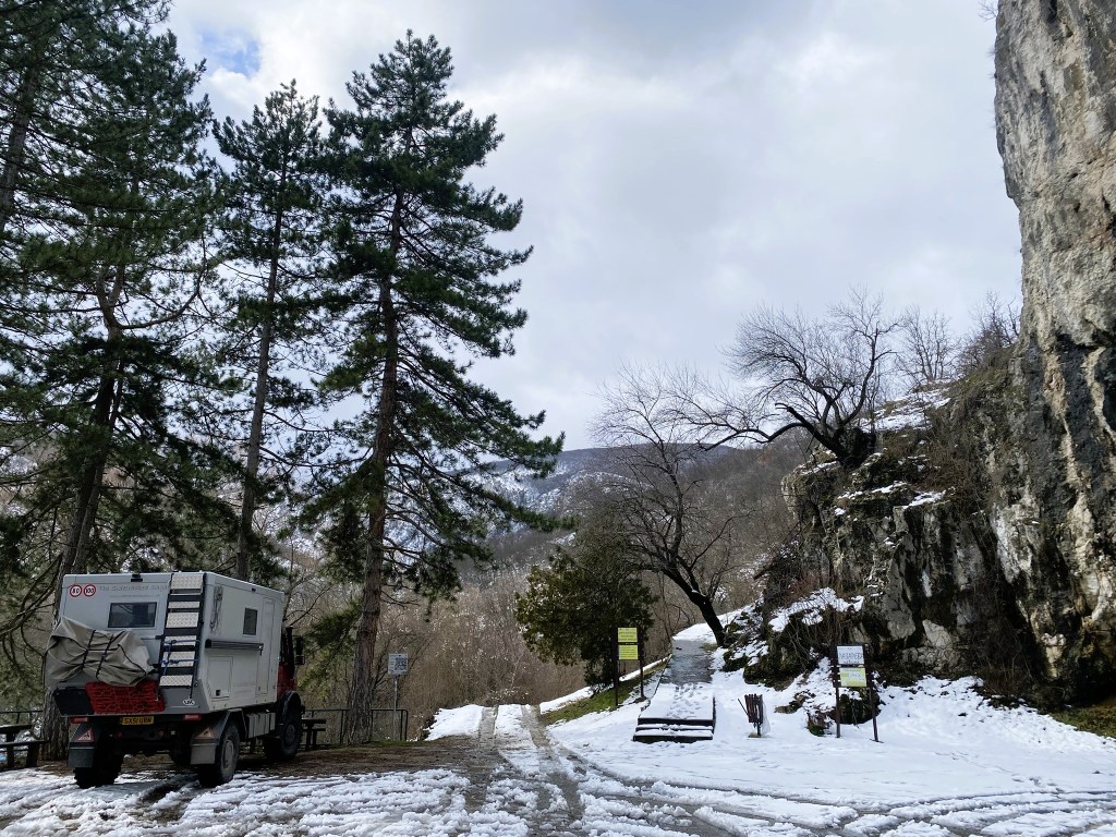 Snow-covered landscape with tall pine trees, an off-road vehicle parked on a gravel path, and rocky hills in the background under a cloudy sky.